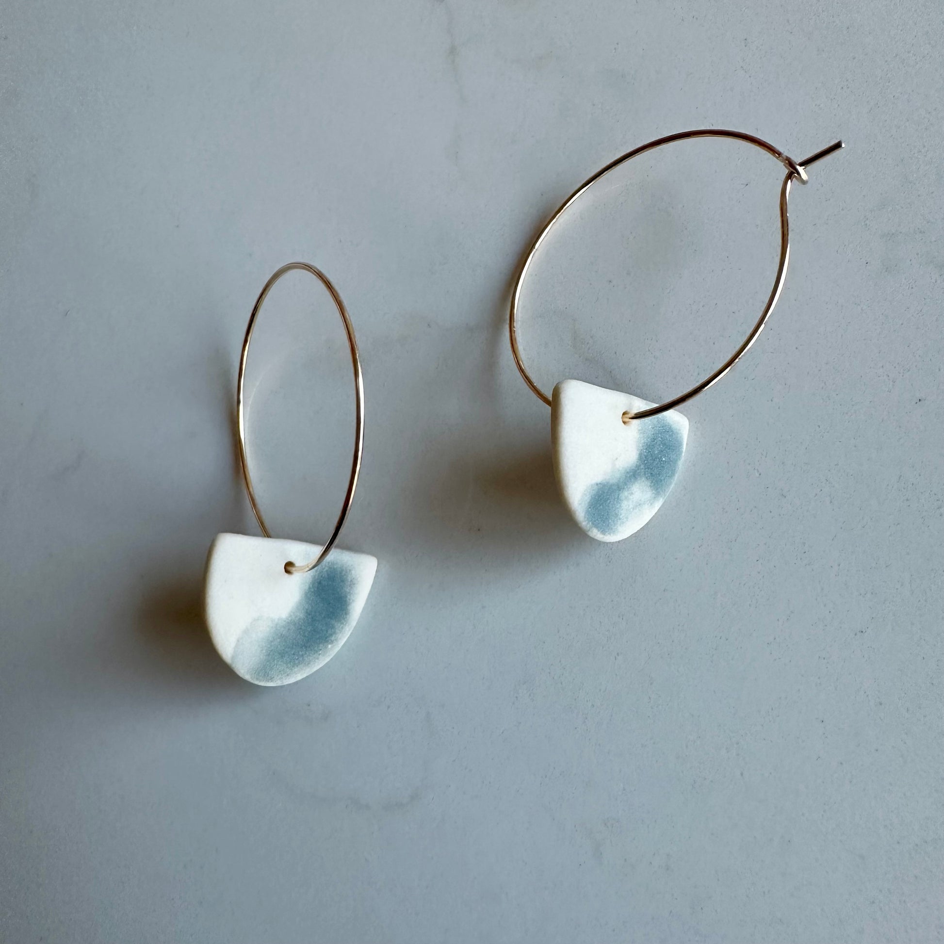 A pair of porcelain earrings with a semi-circular shape and steel underglaze, attached to gold-filled hoops, displayed against a grey background.